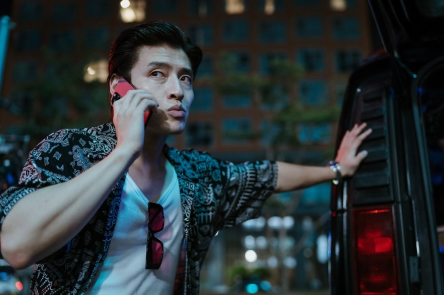 밤에 한 남자가 차 옆에 서서 전화 통화를 하는 모습을 담고 있다. 그는 검은색 패턴 셔츠와 흰색 티셔츠를 입고 있으며, 약간 찡그린 표정으로 통화에 집중하고 있다. 선글라스는 티셔츠에 걸려 있고, 한 손은 차에 기댄 채 서 있다.