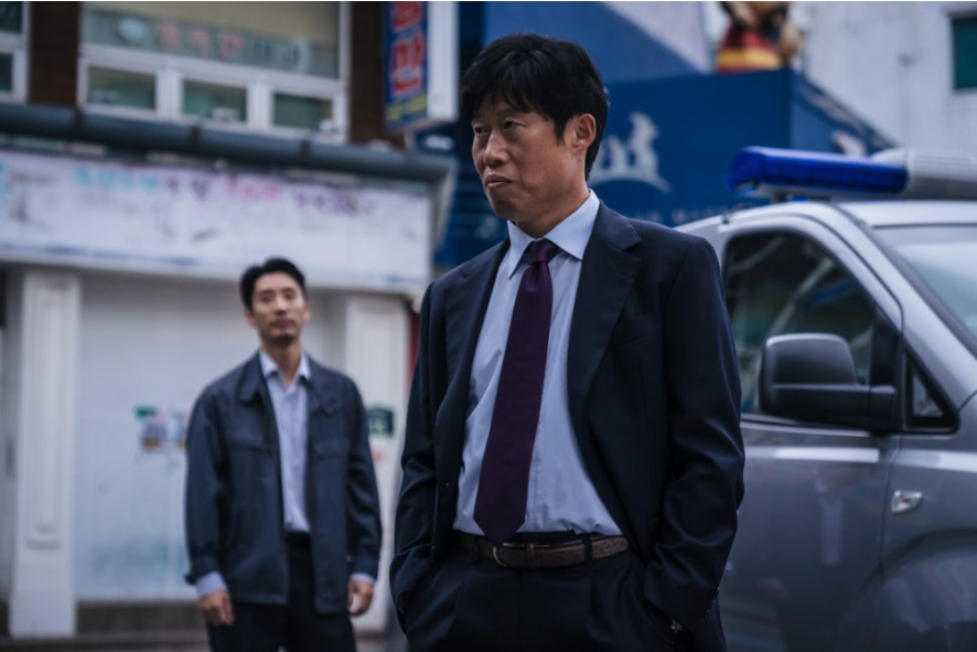 남성 두 명이 서 있는 사진이다. 앞에 있는 남성은 정장을 입고 굳은 표정으로 서 있으며, 손을 바지 주머니에 넣고 있다. 그의 뒤편에는 다른 남성이 캐주얼한 옷차림으로 서 있다. 배경에는 건물과 경찰차로 보이는 차량이 일부 보인다.