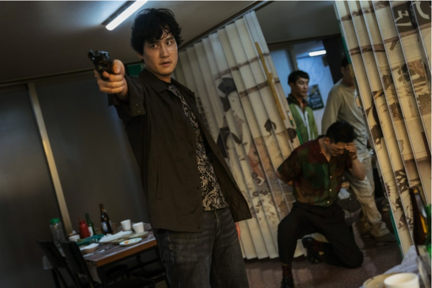 한 남성이 총을 들고 정면을 향해 겨누고 있는 사진이다. 그는 어두운 셔츠를 입고 있으며, 옆으로는 주름진 커튼이 보인다. 그의 시선은 긴장감 넘치는 표정과 함께 목표물을 향하고 있다. 배경에는 다른 두 명의 남성이 서 있거나 무릎을 꿇고 있다.