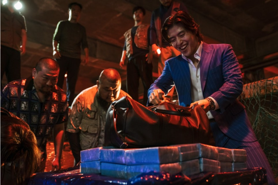 파란색 정장을 입은 한 남성이 환한 미소를 지으며 가방을 열고 있다. 그의 앞에는 파란색 덩어리들이 쌓여 있고, 주변에는 여러 남성들이 그를 지켜보고 있다. 전체적으로 어두운 분위기 속에서 남자의 미소와 파란색 물체가 대비를 이룬다.