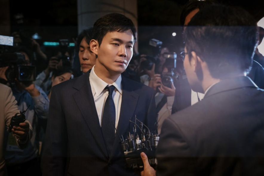 정장을 입은 남성이 수많은 기자들 앞에서 인터뷰를 하는 모습이다. 그는 진지한 표정으로 마이크를 든 사람을 응시하고 있으며, 주변에는 카메라 플래시와 취재진이 가득하다. 사진은 기자회견이나 언론 브리핑 현장의 긴박한 분위기를 담고 있다.