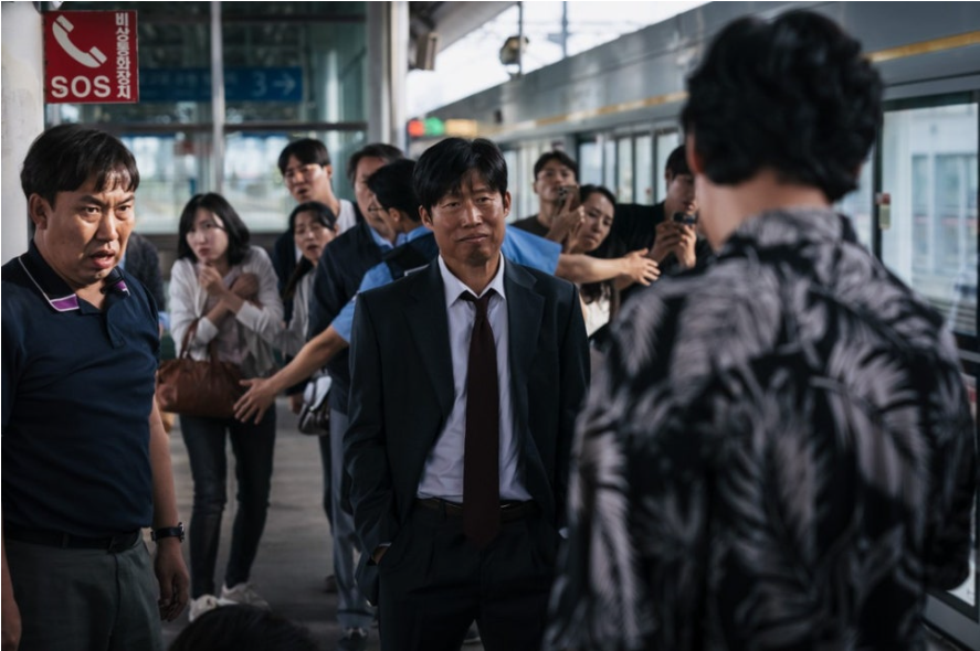 플랫폼에서 두 명의 남성이 서로를 마주 보고 있다. 한 남성은 정장을 입고 굳은 표정으로 서 있으며, 다른 남성은 패턴 셔츠를 입은 채 등을 보이고 있다. 주변에는 겁에 질린 표정의 사람들이 그들을 지켜보고 있어 긴장감을 조성한다.