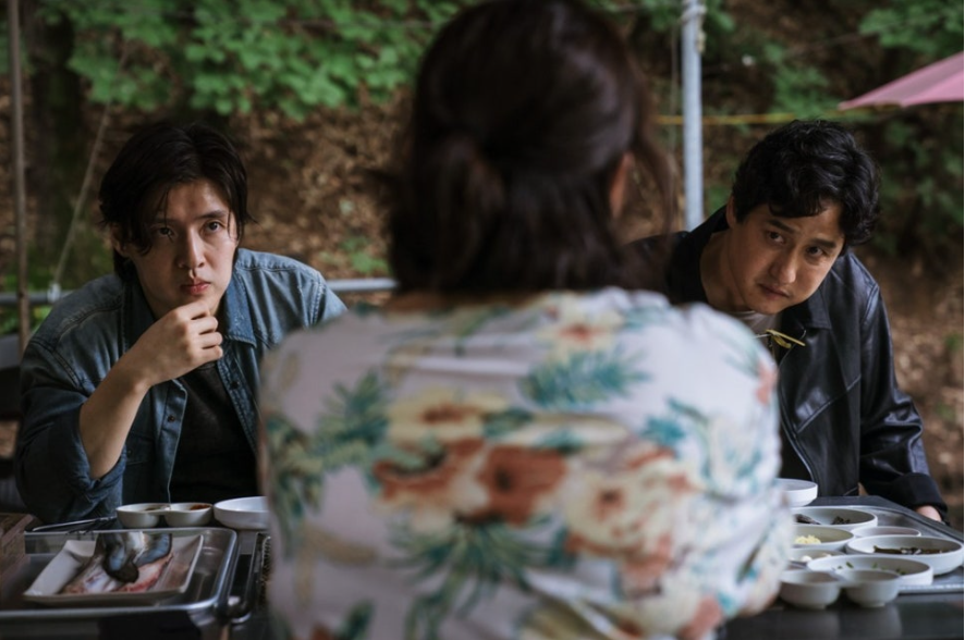 세 명이 테이블에 둘러앉아 있는 모습이다. 뒷모습이 보이는 여성을 중심으로 두 남성이 마주 보고 앉아 있다. 왼쪽에 데님 재킷을 입은 남성은 심각한 표정이고, 오른쪽에 가죽 재킷을 입은 남성은 무언가 걱정하는 듯한 표정을 짓고 있다.