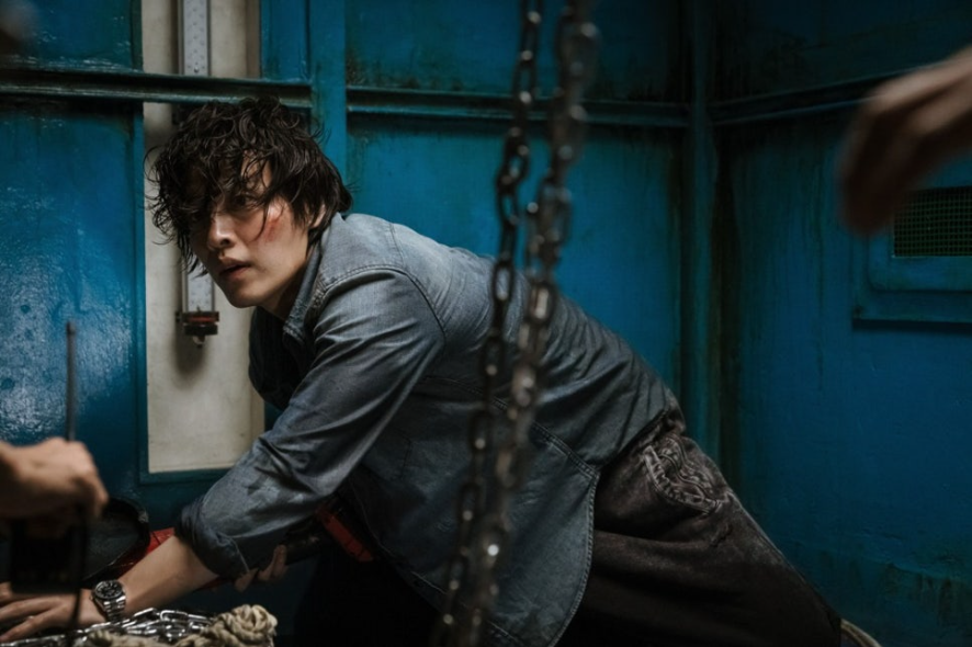 낡은 푸른색 방 안에서 한 남성이 쇠사슬에 묶인 채 바닥에 웅크리고 있다. 그의 얼굴에는 상처가 있고, 옷은 더러워져 있다. 혼란스러워 보이는 표정으로 정면을 응시하며, 긴박하고 절박한 상황에 놓여 있음을 보여준다.