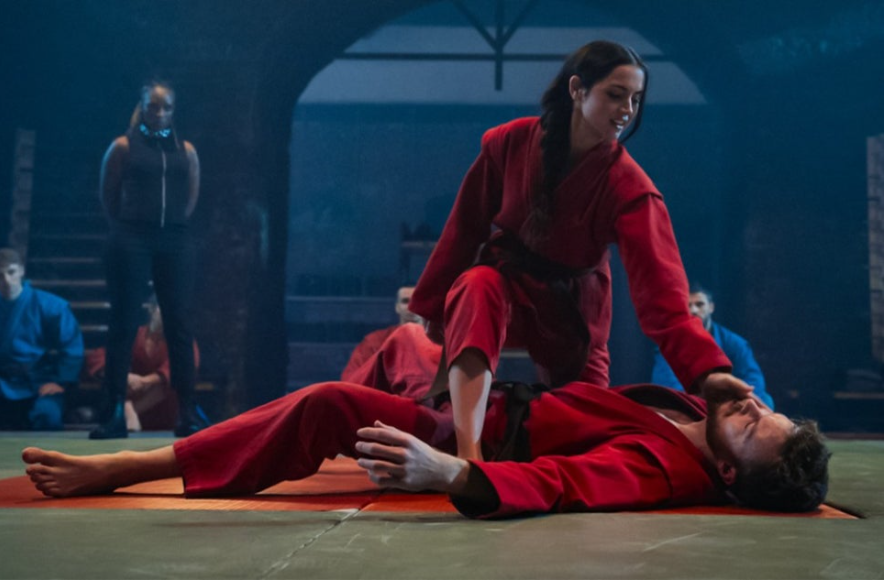 이미지는 영화 **《발레리나》**의 스틸컷으로, 주인공인 루니(아나 데 아르마스)가 훈련을 하는 장면입니다. 
붉은색 유도복을 입은 루니가 다른 사람을 제압하고 있으며, 배경에는 다른 훈련생들이 앉아 있습니다. 이 장면은 주인공이 숙련된 암살자로 성장하는 과정을 보여주는 훈련 시퀀스 중 일부로 보입니다.