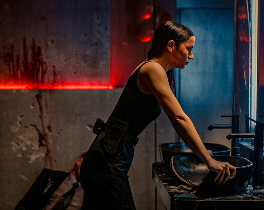 이미지는 영화 **《발레리나》**의 스틸컷입니다. 주인공인 루니(아나 데 아르마스)가 화장실로 보이는 공간에서 거울을 보며 세면대에 몸을 기댄 채 숨을 고르는 모습입니다. 벽에는 핏자국이 선명하고, 바닥에는 쓰러져 있는 사람이 있어 방금 치열한 전투를 벌였음을 암시합니다.