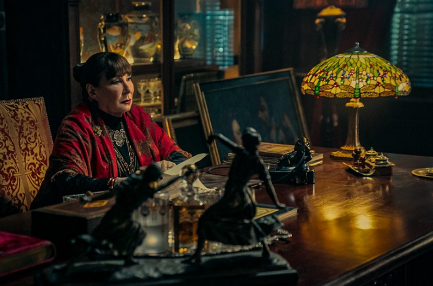 이미지는 영화 **《발레리나》**의 스틸컷으로, 안젤리카 휴스턴이 연기한 디렉터가 책상에 앉아 있는 모습입니다. 그녀의 앞에는 두 명의 발레리나 조각상이 놓여 있어 '발레리나' 집단의 수장임을 암시합니다. 주변의 고풍스러운 가구와 조명은 그녀의 우아하면서도 위압적인 분위기를 강조합니다.