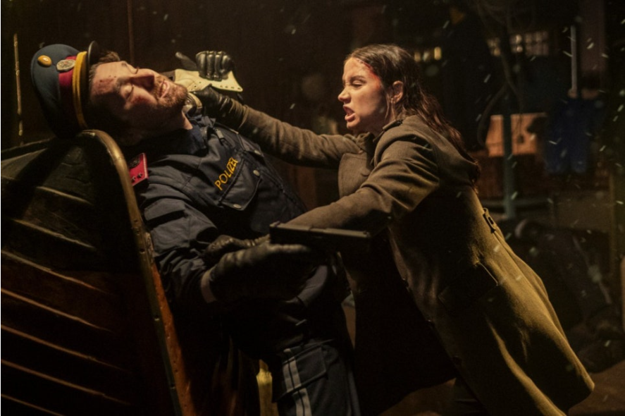 이미지는 영화 **《발레리나》**의 스틸컷입니다. 주인공인 루니(아나 데 아르마스)가 제복을 입은 남성과 격렬한 몸싸움을 벌이는 장면을 담고 있습니다. 그녀의 얼굴에는 피가 묻어 있고 표정은 매우 분노에 차 있으며, 손에는 총을 쥐고 있어 긴박한 전투 상황임을 알 수 있습니다.