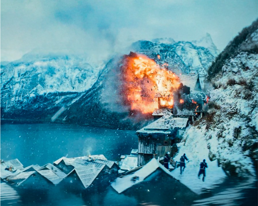 이미지는 영화 **《발레리나》**의 스틸컷입니다. 눈 덮인 산과 호수가 있는 겨울 풍경 속에서, 건물 한 채가 폭발하는 장면을 담고 있습니다. 불꽃과 연기가 강렬하게 솟아오르고 있으며, 아래에는 무장한 사람들이 뛰어가는 모습이 보여 영화 속 거대한 액션과 파괴적인 스케일을 짐작하게 합니다.