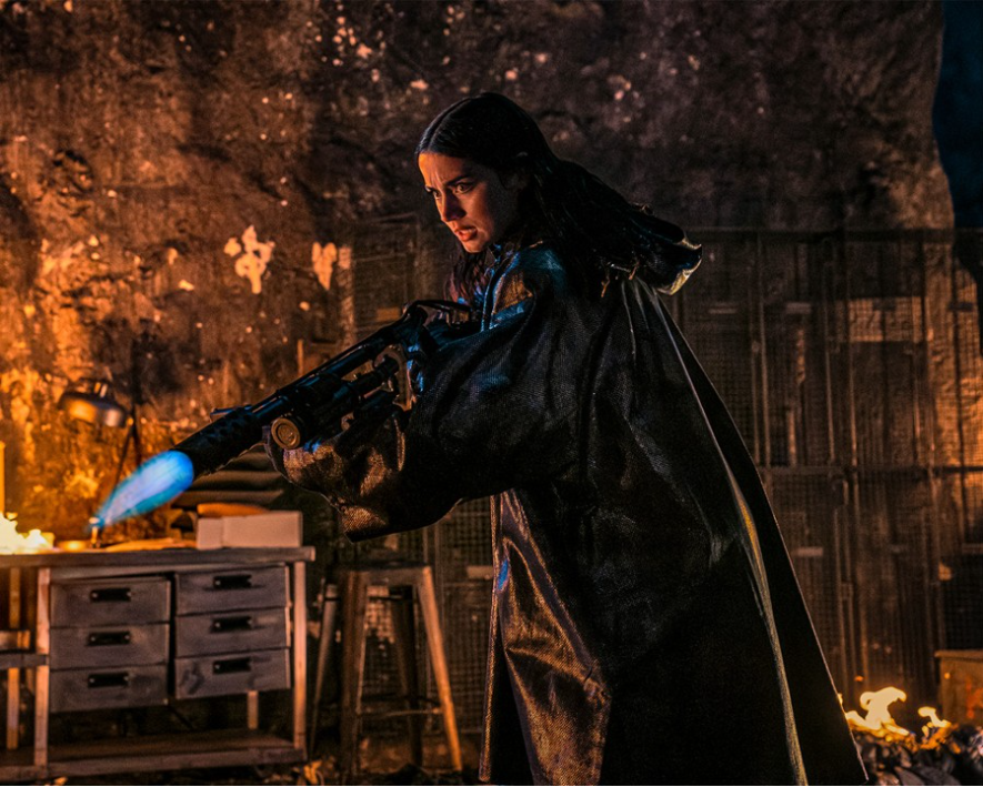 이미지는 영화 **《발레리나》**의 스틸컷입니다. 주인공인 루니(아나 데 아르마스)가 검은 망토를 두르고 화염 방사기를 들고 있는 모습입니다. 주변은 어둡고 불이 붙어 있어 긴박한 상황임을 암시하며, 그녀의 비장한 표정에서 복수를 향한 강한 의지를 느낄 수 있습니다.