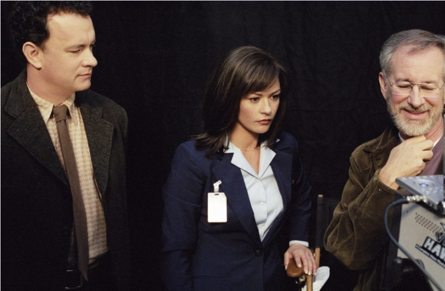 영화 터미널의 배우와 감독이 함께 있습니다. 왼쪽부터 주연 배우 톰 행크스, 캐서린 제타존스, 그리고 스티븐 스필버그 감독입니다. 이들은 촬영 현장에서 무언가를 주의 깊게 바라보고 있습니다.