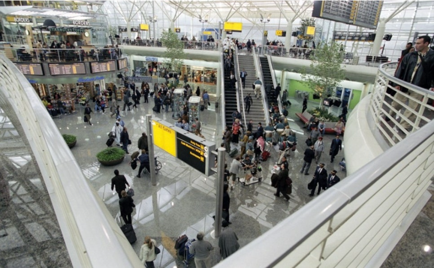 공항 터미널 내부를 위에서 내려다본 모습입니다. 사람들로 붐비는 1층에는 여러 상점과 안내 표지판이 보이고, 에스컬레이터를 이용해 위층으로 올라가는 사람들도 있습니다. 높은 천장과 현대적인 디자인의 난간이 눈에 띕니다.