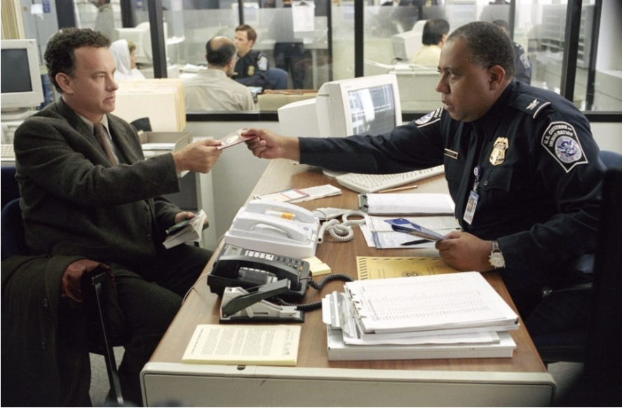 이 사진은 영화 '터미널'의 한 장면입니다. 톰 행크스가 공항 세관원에게 여권을 건네고 있습니다. 책상 위에는 서류와 전화기들이 어지럽게 놓여 있으며, 뒤쪽으로는 다른 직원들이 근무하는 사무실 공간이 보입니다.