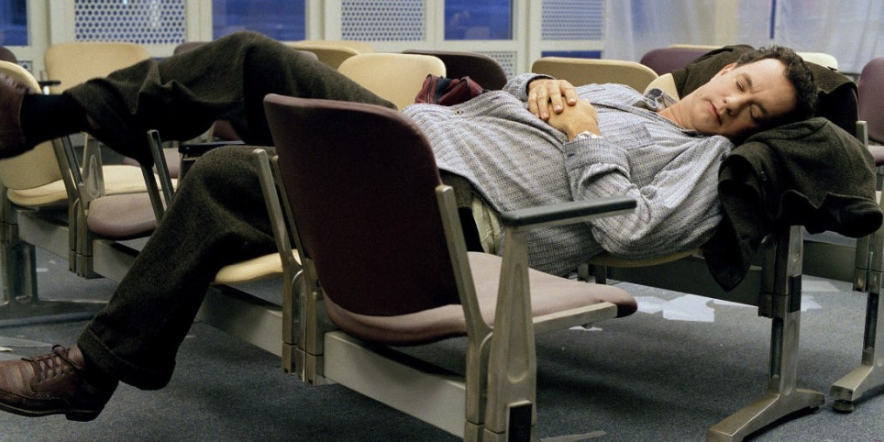 이 사진은 영화 '터미널'의 주인공 톰 행크스가 공항 의자 여러 개에 걸쳐 잠들어 있는 모습을 보여줍니다. 여러 날 공항에 머물면서 지쳐 잠든 그의 고단한 상황을 잘 보여주는 장면입니다.