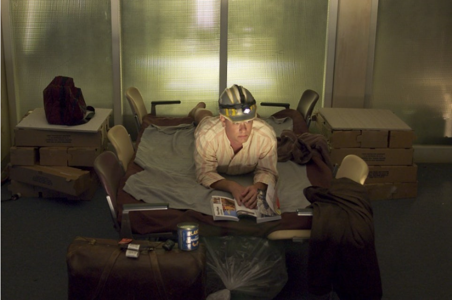 이 사진은 영화 '터미널'에서 주인공인 톰 행크스가 공항에서 생활하는 모습을 보여줍니다. 그는 여러 개의 의자를 이어붙여 침대 삼아 누워있고, 머리에는 헤드랜턴을 쓰고 잡지를 읽고 있습니다. 그의 옆에는 서류 가방과 통조림 캔, 쓰레기봉투 등이 놓여있어 공항이 그의 임시 거주지가 되었음을 알 수 있습니다.