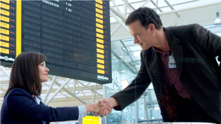 이 사진은 영화 '터미널'의 한 장면으로, 톰 행크스와 캐서린 제타존스가 공항에서 악수를 나누는 모습을 보여줍니다. 두 사람은 서로 마주 보며 미소 짓고 있으며, 뒤로는 항공편 정보가 표시된 전광판이 보입니다.