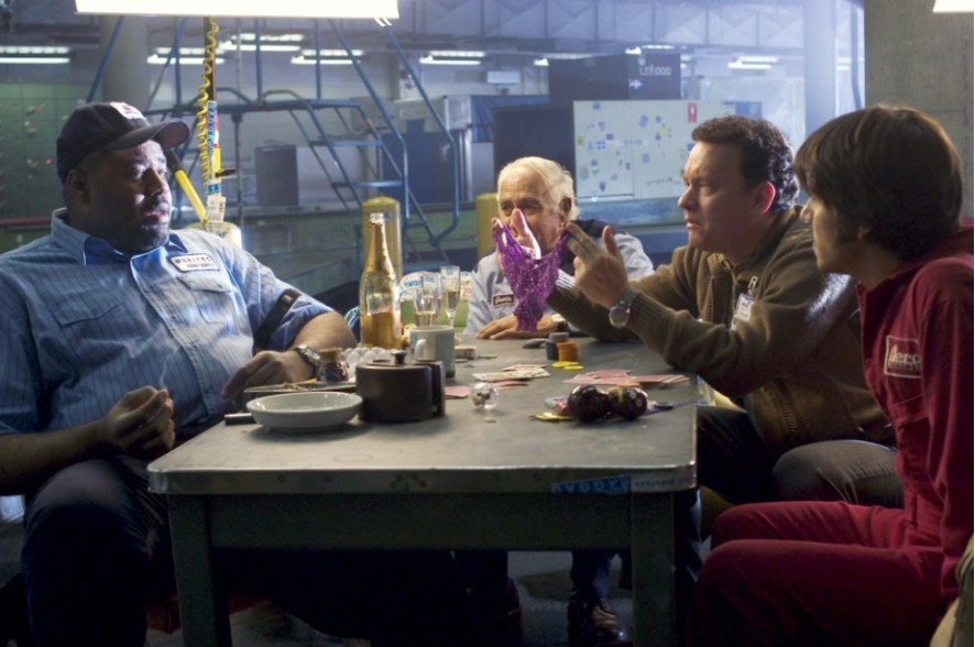 이 사진은 영화 '터미널'에서 주인공인 톰 행크스가 공항 직원들과 함께 둘러앉아 카드 게임을 하는 장면입니다. 이들은 공항의 후미진 공간에 탁자를 두고 앉아 함께 시간을 보내고 있습니다. 톰 행크스가 다른 사람에게 무언가 설명하듯 손짓하고 있어, 이들이 공항에서 함께 동료애를 쌓아가는 모습을 보여줍니다.