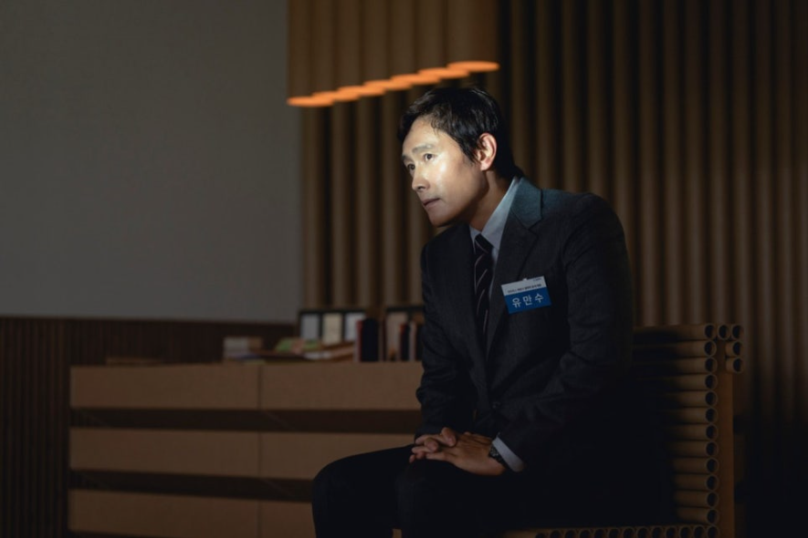 유만수는 깔끔한 정장 차림에 명찰을 달고 어두운 실내에서 긴장한 듯 앉아 있는 모습으로, 재취업을 위해 고군분투하는 캐릭터의 상황을 보여줍니다.