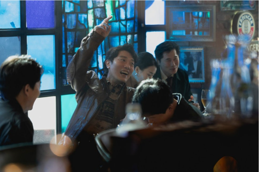 가운데 남성(박희순)은 웃으며 한 손을 높이 들고 있으며, 주변 사람들은 그를 바라보며 함께 미소 짓고 있습니다. 활기차고 흥겨운 순간을 보여줍니다.