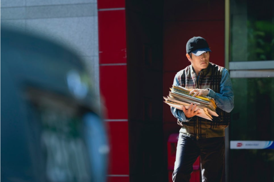 이 스틸컷은 영화 어쩔 수가 없다의 주인공 유만수(이병헌)가 모자와 캐주얼한 복장으로 무언가 잔뜩 쌓인 서류 뭉치를 품에 안고 경계하는 듯한 표정으로 주위를 살피는 모습을 담고 있습니다. 그의 절박한 상황이 느껴집니다.