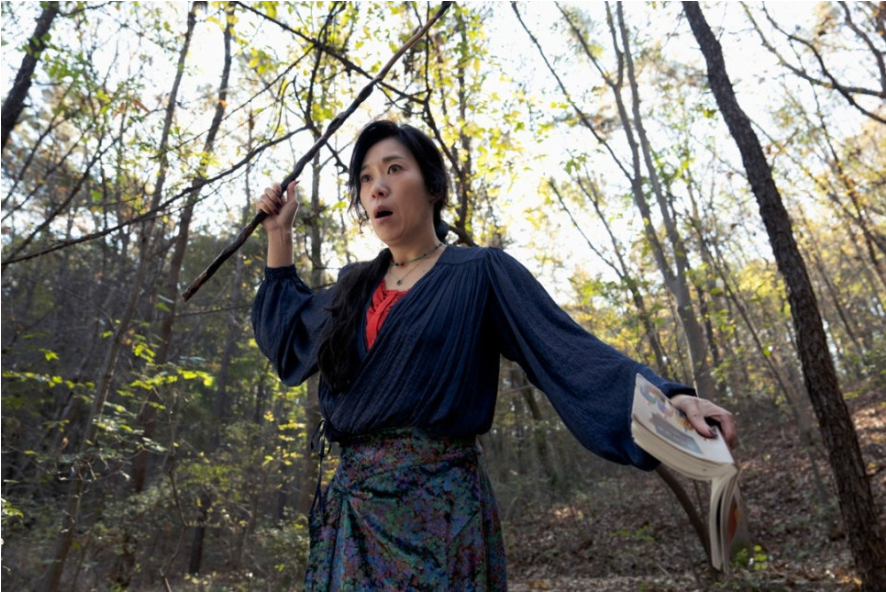 이 스틸컷은 영화 어쩔 수가 없다에서 이아라(염혜란)가 숲속에서 지팡이와 책을 든 채 놀란 표정으로 위를 올려다보는 모습을 담고 있습니다.