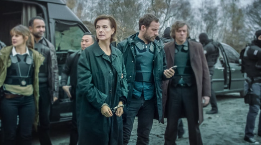 수갑을 찬 중년 여성(연쇄살인범 '사마귀')이 어두운 야외에서 형사들에게 둘러싸여 이동하는 장면입니다. 주변에는 다른 형사들 혹은 무장한 인물들이 보이며, 긴장감 넘치는 분위기를 보여줍니다. 여성은 검은색 코트를 입고 무표정한 얼굴입니다.