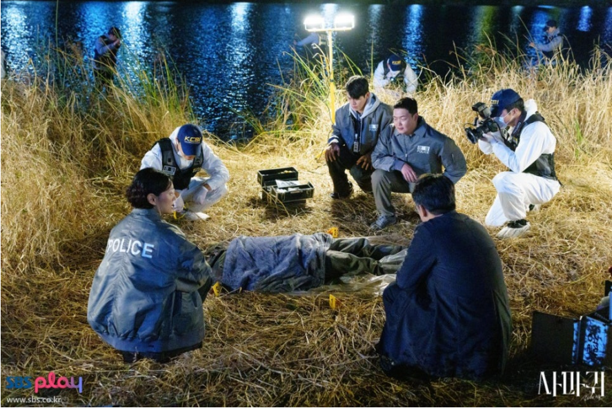 밤 시간, 물가 갈대밭에서 경찰과 국과수 요원들이 변사체를 둘러싸고 현장을 조사하는 모습입니다. 중앙에 시신이 담요로 덮여 있으며, 주변에는 경찰(POLICE)과 과학수사요원(KCSI) 복장의 사람들이 사건의 단서를 찾는 긴박한 분위기를 보여줍니다.