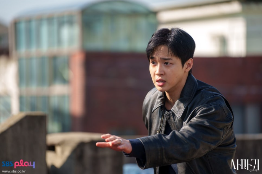 배우 장동윤으로 보이는 젊은 남성 배우가 가죽 재킷을 입고 어딘가를 향해 손을 내밀며 놀라거나 절박한 표정을 짓고 있습니다. 야외 건물 옥상 같은 곳에서 촬영되었으며, 그의 긴장된 표정이 극의 긴박한 상황을 암시합니다.