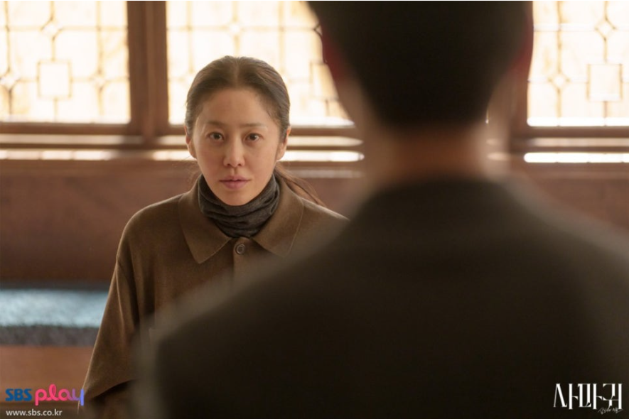 배우 고현정으로 보이는 여주인공이 갈색 셔츠형 외투와 터틀넥을 입고 정면을 응시하고 있습니다. 그녀는 심각하고 무거운 표정을 짓고 있으며, 전경에는 뒷모습을 보인 남성 캐릭터의 어깨가 클로즈업되어 있어 두 인물 간의 긴장된 대화 상황을 암시합니다.