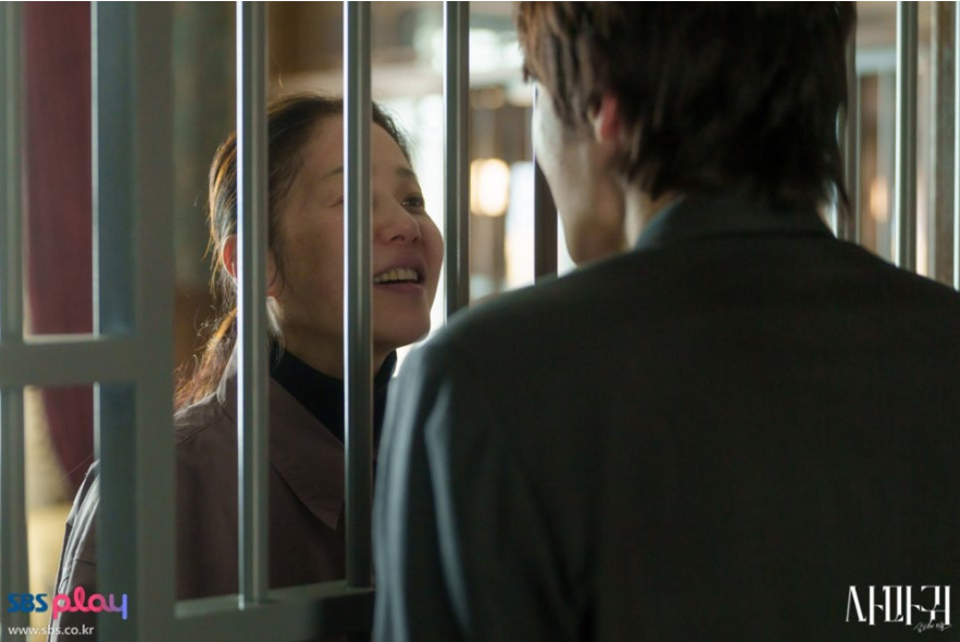 배우 고현정으로 보이는 여주인공이 쇠창살 안에서 바깥의 남성을 바라보며 미소를 짓는 모습입니다. 이전 스틸컷들과 달리 밝고 해맑은 표정을 하고 있으며, 전경에는 남성의 뒷모습이 클로즈업되어 있어 두 인물의 특별한 관계를 암시합니다.