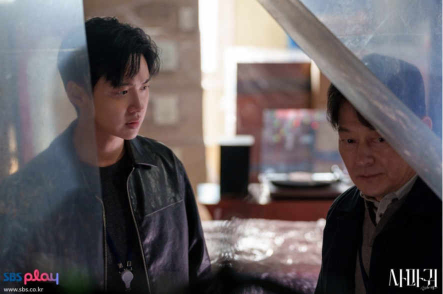 배우 장동윤과 또 다른 중년 남성 배우 조성하가 실내에서 함께 있는 장면입니다. 장동윤은 가죽 재킷을 입고 심각한 표정으로 정면을 응시하고 있으며, 중년 남성 조성하는 옆에서 고개를 숙이고 있습니다. 이들은 투명한 막 너머에서 포착되어 긴장감을 더합니다.