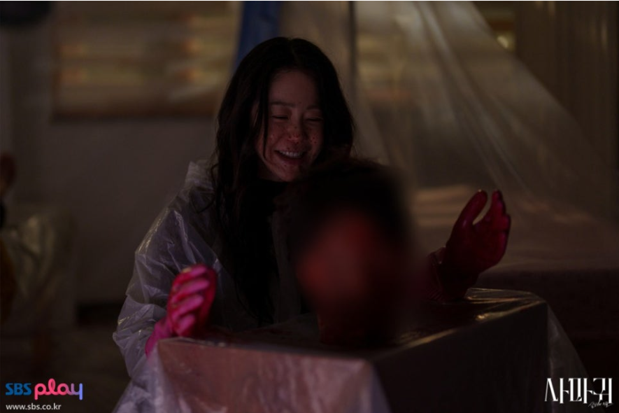 배우 고현정으로 보이는 여주인공이 방진복과 붉은색 장갑을 낀 채 어두운 실내에서 크게 웃고 있는 충격적인 장면입니다. 그녀의 앞에는 블러 처리된 무언가가 놓여 있으며, 이 섬뜩한 미소는 극 중 캐릭터의 광기를 암시하며 강한 긴장감을 조성합니다.